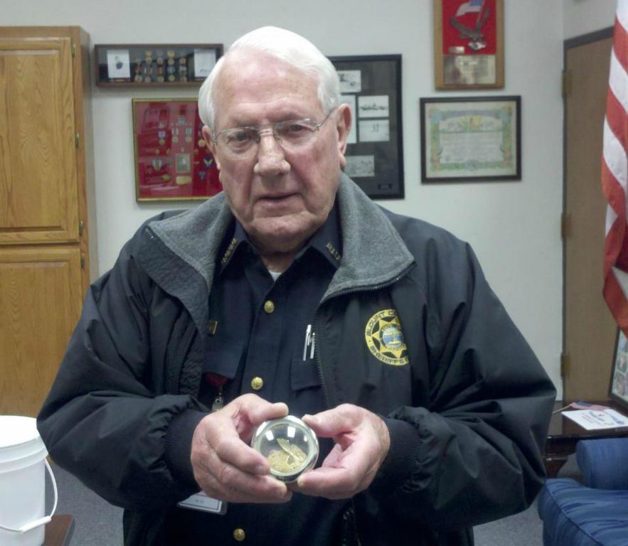 Man in glasses and black Sheriff's office coat holds up circular item and looks into the camera